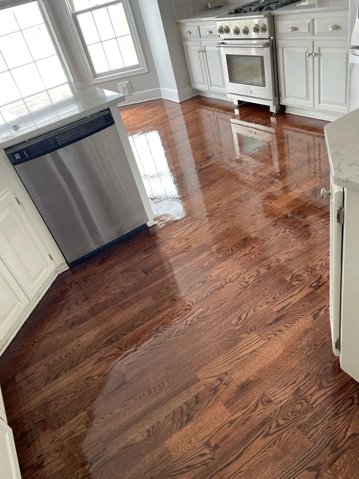 Refinished dark oak kitchen floor in front of a stainless dishwasher and white cabinets with window light across the boards, in Leawood, Kansas